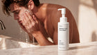 A bottle of ARNEUX Blemish Care Face Wash stands on a marble countertop in a bathroom, with a man washing his face in the background