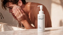 A bottle of ARNEUX Blemish Care Face Wash stands on a marble countertop in a bathroom, with a man washing his face in the background