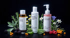 Three ARNEUX skincare bottles arranged on a black background with botanical ingredients around them: a clear bottle of BiPhasic Makeup Remover with amber liquid, a white bottle of Micellar Cleansing Water, and a pink pump bottle of Radiant Glow Facial Wash
