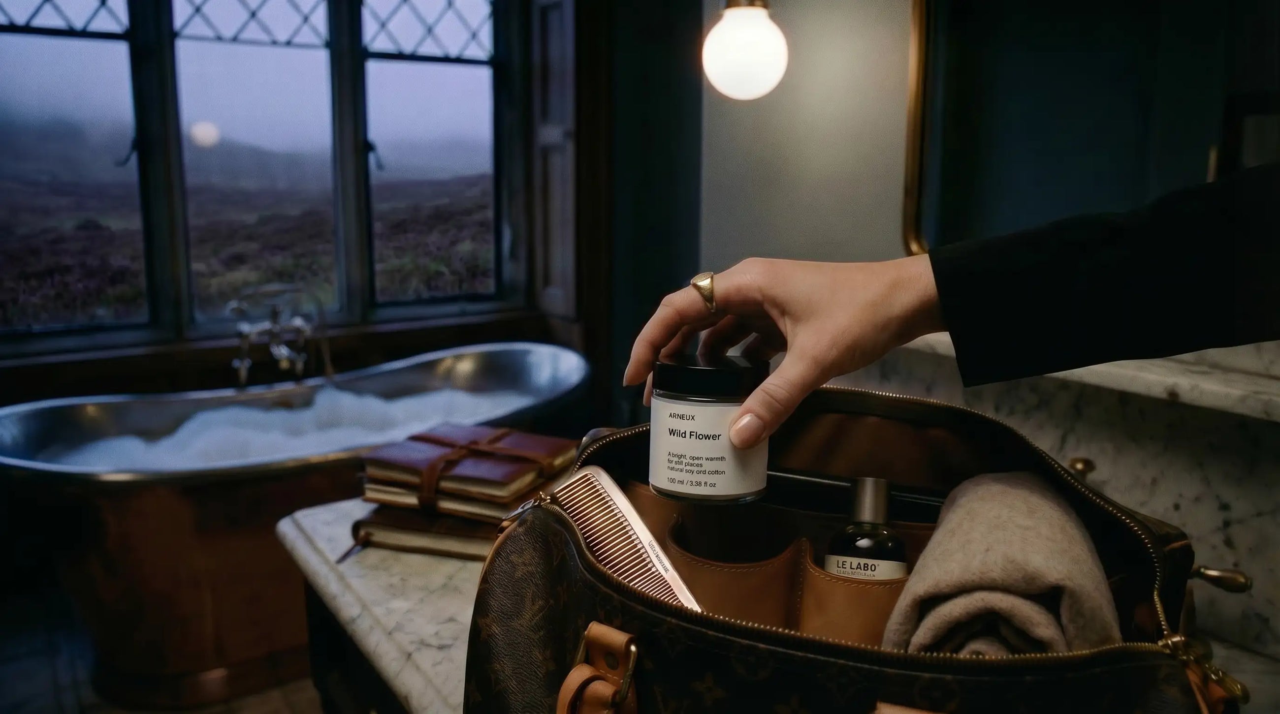 Woman reaching for WILD FLOWER travel candle in luxury travel bag, moody highland inn interior at dusk
