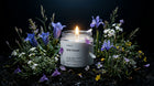 WILD FLOWER travel candle burning among purple iris, white blossoms and wild meadow grasses on dark background