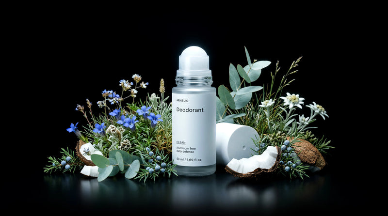 A ARNEUX roll-on deodorant bottle with its cap off, styled with green foliage, blue wildflowers, and coconut halves on a reflective black surface against a black background