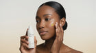 Woman dotting AURA vitamin C serum onto cheekbone with fingertip, bright even skin tone close-up