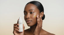 Woman dotting AURA vitamin C serum onto cheekbone with fingertip, bright even skin tone close-up